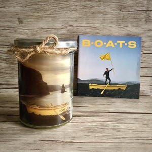 Op de afbeelding: Een glazen pot met een kaars met een foto van een boot met het woord "HOME" erop. De kaars is vastgebonden met touw en staat naast een CD met de titel "B.O.A.T.S."