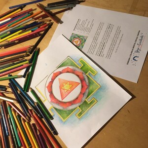 May include: A drawing of a mandala with a red triangle in the center, surrounded by a green square and a blue circle. The drawing is on a white sheet of paper and there are colored pencils on the table.
