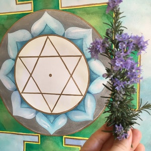 Può includere: Un dipinto ad acquerello di un mandala verde e oro con un disegno di fiore di loto blu. Una mano tiene un ramo di fiori di lavanda.