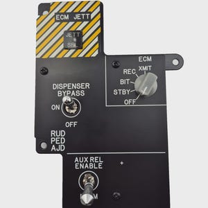 Puede incluir: Un panel de control negro con varios interruptores y etiquetas. El panel presenta texto como "ECM JETT", "DISPENSER BYPASS" y "AUX REL ENABLE". Tiene una sección a rayas amarillas y blancas y múltiples perillas e interruptores.