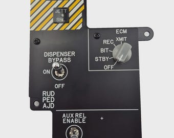 May include: A black control panel with various switches and labels. The panel features text such as "ECM JETT", "DISPENSER BYPASS", and "AUX REL ENABLE". It has a yellow and white striped section and multiple knobs and toggle switches.
