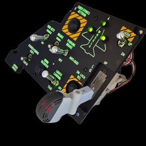 Puede incluir: Un panel de control de avión negro con múltiples interruptores de palanca, botones y luces indicadoras verdes. El panel incluye etiquetas como EMER STORES JETTISON, PARKING BRAKE y HOOK, con una palanca gris unida a la sección inferior.