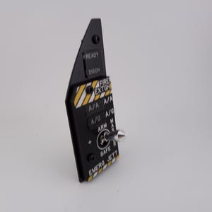 Puede incluir: Panel negro con una variedad de interruptores y etiquetas, incluyendo "READY", "DISCH", "FIRE EXTGH", "A/A", "A/G", "ARM", "SAFE", "EMERG JETT".