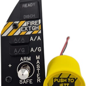 Op de afbeelding: Een zwart bedieningspaneel met witte tekst READY, DISCH, FIRE EXTG, A/A, A/G, ARM, MASTER, SAFE en EMERG JETT, voorzien van gele en zwarte waarschuwingsstrepen. Daarnaast staat een cilindervormige gele knop met het opschrift PUSH TO JETT en strepen.
