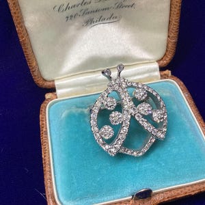 May include: A silver-toned ladybug brooch encrusted with clear, sparkling rhinestones. The insect-shaped pin features a rounded body with circular patterns and two small antennae at the top, presented inside a vintage-style jewelry box.
