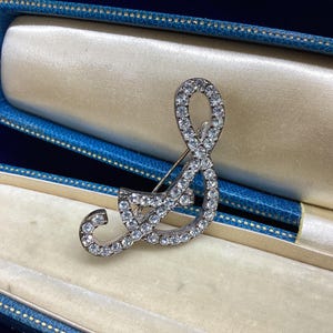 May include: A silver-toned brooch in the shape of a musical treble clef, encrusted with clear, sparkling gemstones. The brooch is displayed inside a vintage blue and gold jewelry box, with a soft, cream-colored interior.