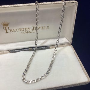 May include: A silver-toned mariner link chain necklace displayed inside a rectangular jewelry box. The interior of the box features a white satin lining and the printed text Precious Jewels by Milano with a crown icon above the brand name.