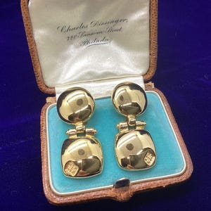 May include: A pair of gold-toned drop earrings featuring a circular top piece connected by a hinge to a square bottom piece. The earrings are displayed inside a vintage brown jewelry box with a light blue interior and the text Charles Diesinger, 720 Sansom Street, Philada printed on the inside lid.