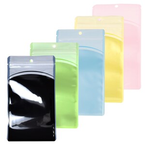 Op de afbeelding: Vijf transparante, hersluitbare plastic zakjes met een ophanggat bovenaan. De zakjes zijn in verschillende kleuren: zwart, groen, lichtblauw, geel en roze.