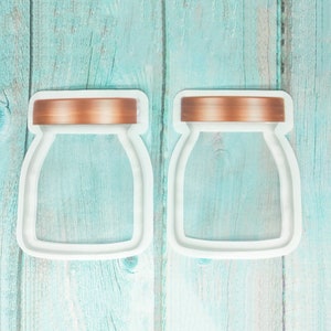 May include: Two white cookie cutters shaped like jars with a copper-colored lid. The cookie cutters are on a light blue wooden background.