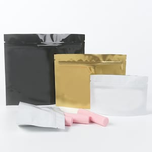 Puede incluir: Tres bolsas de pie de diferentes colores con cierre de cremallera. Las bolsas son negras, doradas y plateadas. Hay cuatro tubos de bálsamo labial rosa delante de las bolsas.