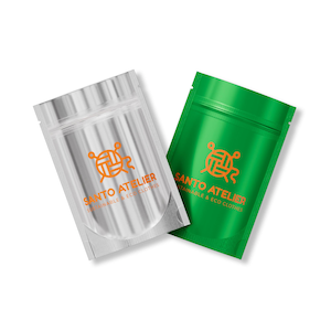 May include: Two small, sealed pouches. One is silver, the other is bright green. Both have an orange logo and the text "SANTO ATELIER SUSTAINABLE & ECO CLOTHES" printed on them. The pouches are likely for packaging.