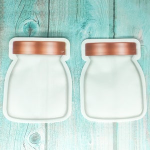 May include: Two white reusable food storage bags with copper lids. The bags are shaped like jars and are laying flat on a teal wooden surface.