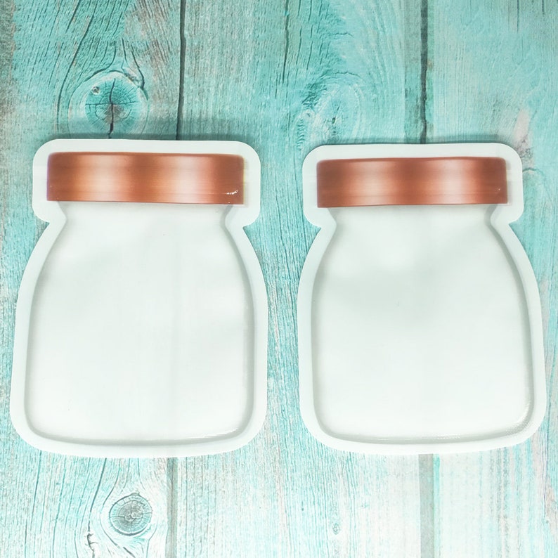 May include: Two white reusable food storage bags with copper lids. The bags are shaped like jars and are laying flat on a teal wooden surface.