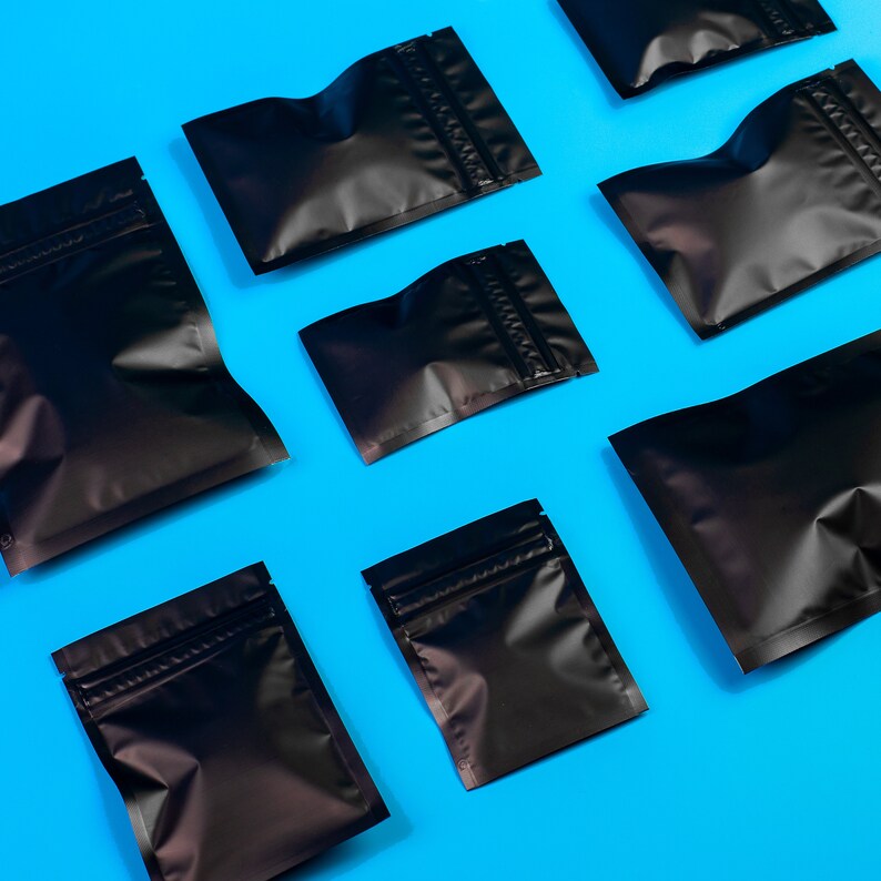 May include: A flat lay of nine black zippered pouches on a bright blue background. The pouches are various sizes and are arranged in a staggered pattern.