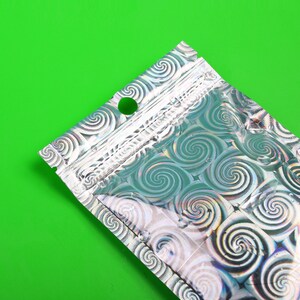 May include: A silver, holographic bag with a swirl pattern. The bag has a small hole near the top and a zip closure. The background is a solid, bright green.