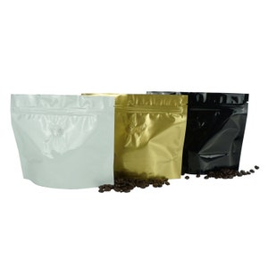 May include: Three stand-up pouches with zipper closures. The pouches are made of a shiny, metallic material. The pouches are white, gold, and black. Coffee beans are scattered in front of the pouches.