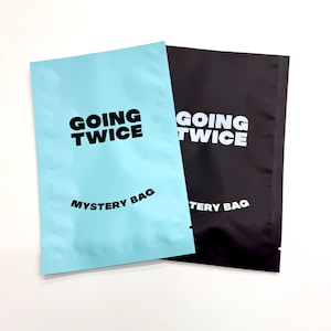 May include: Two mystery bags are shown, one in light blue and the other in black. The light blue bag has the words "GOING TWICE" and "MYSTERY BAG" printed in black. The black bag has the same text in white, against a white background.