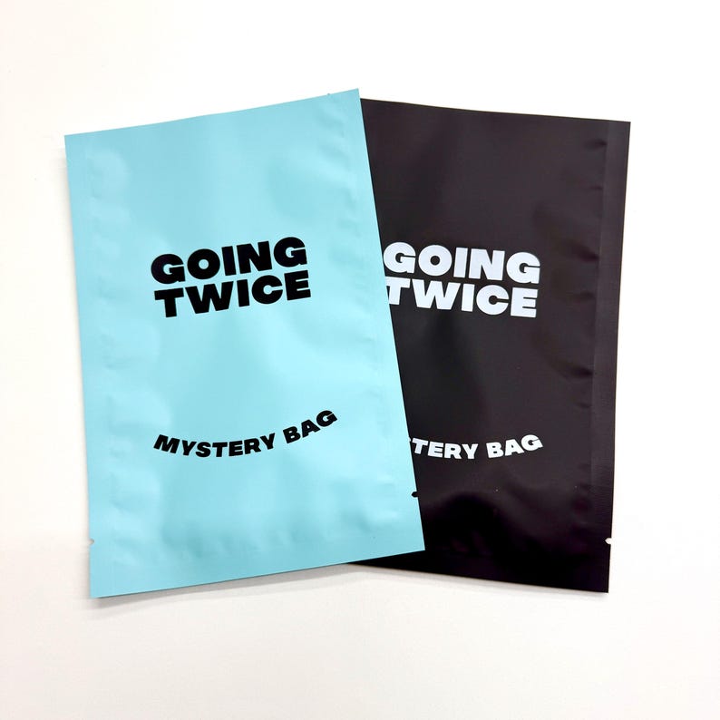 May include: Two mystery bags are shown, one in light blue and the other in black. The light blue bag has the words "GOING TWICE" and "MYSTERY BAG" printed in black. The black bag has the same text in white, against a white background.
