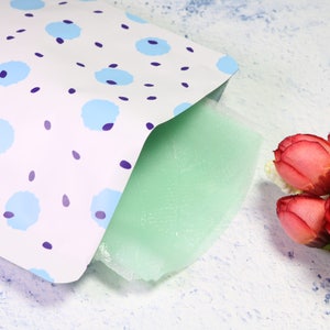 May include: A white pouch with a blue and purple floral pattern, partially open to reveal a green sheet mask.