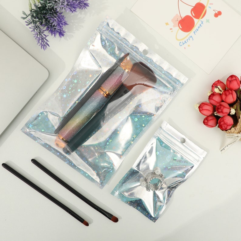 May include: Clear, iridescent zip-top bags containing makeup brushes and a ring. The bags have a holographic effect with star-shaped glitter. Two black makeup brushes are also visible. A card with cherries and the words "Cherry planet" is in the background.