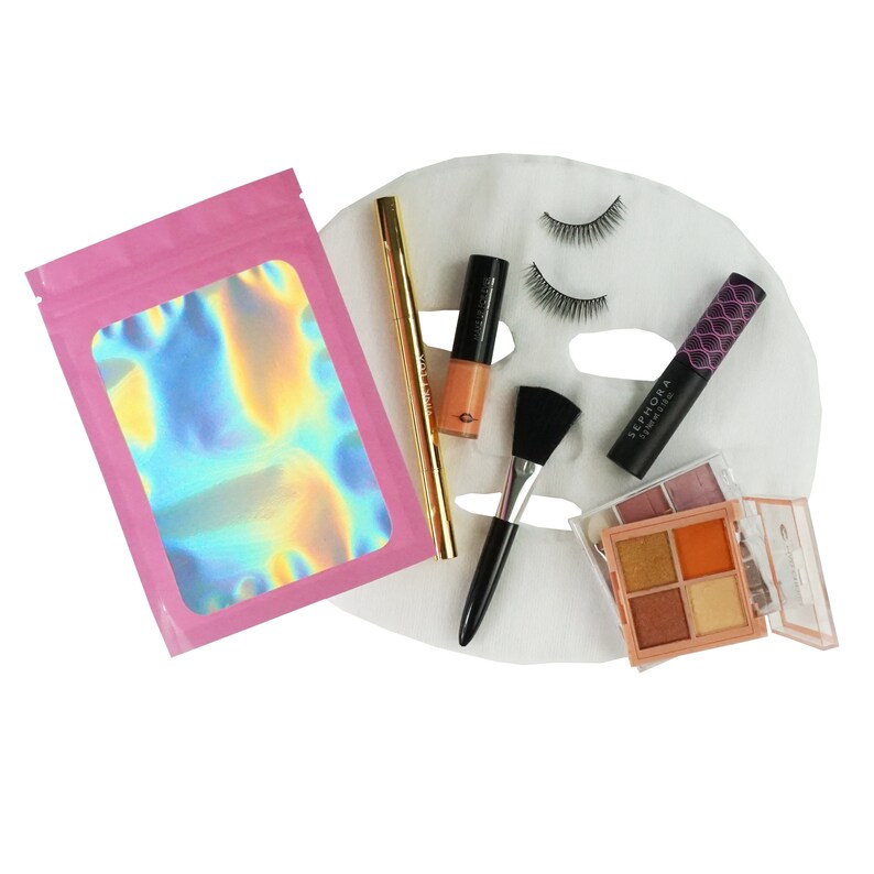 May include: A pink pouch with a holographic iridescent design, a gold eyeliner, a black mascara, a black and gold eyeshadow palette, a black brush, and a white face mask with false eyelashes.