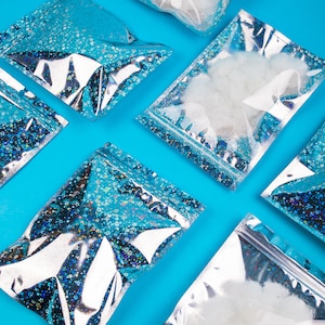 May include: An overhead shot of several clear, resealable bags filled with white fluff and iridescent glitter. The bags are arranged diagonally on a bright blue background. The bags have a silver and blue holographic design with a zipper closure.