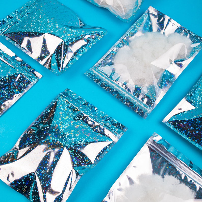 May include: An overhead shot of several clear, resealable bags filled with white fluff and iridescent glitter. The bags are arranged diagonally on a bright blue background. The bags have a silver and blue holographic design with a zipper closure.
