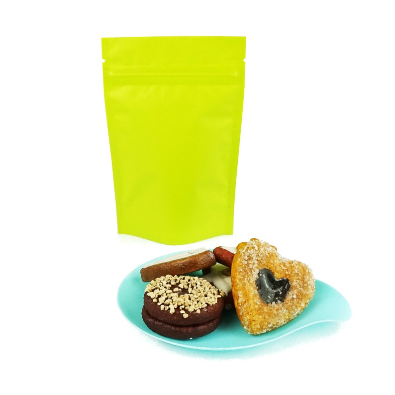 May include: A bright green resealable pouch with a variety of cookies on a blue plate. The cookies include a heart-shaped cookie with a black center, a round cookie with white frosting, and a round cookie with brown frosting and sprinkles.