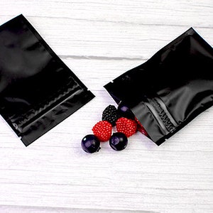 May include: Two black zippered pouches with a zipper closure. The pouches are lying on a white wooden surface. There are red and black candies scattered on the surface.