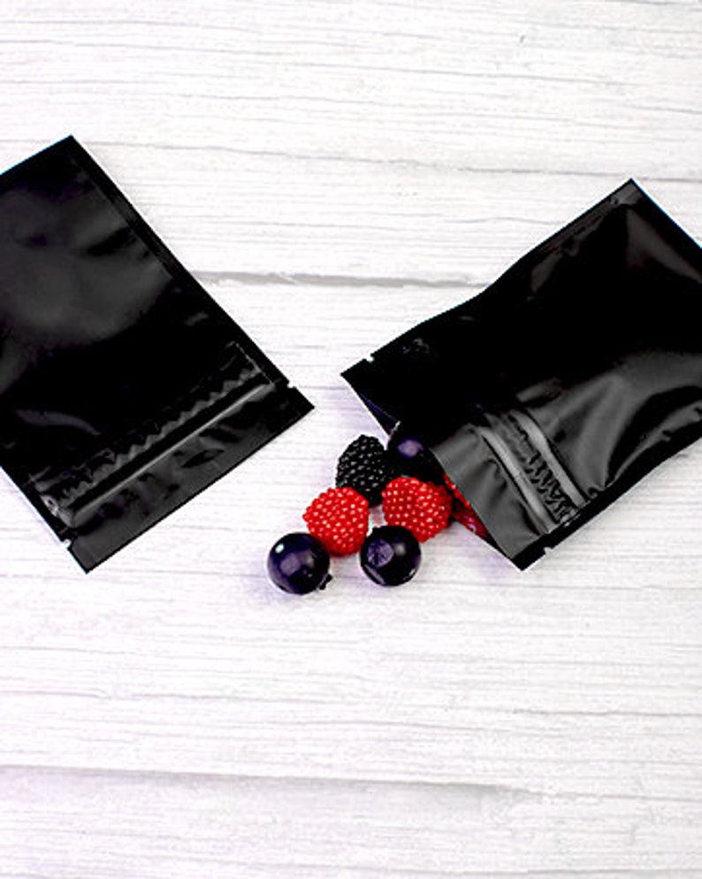 May include: Two black zippered pouches with a zipper closure. The pouches are lying on a white wooden surface. There are red and black candies scattered on the surface.