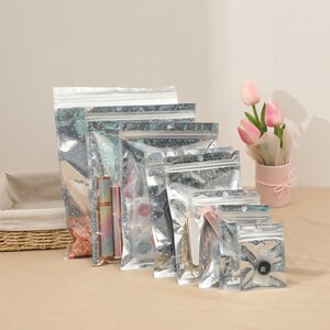 May include: A collection of clear, glittery zip-top bags in various sizes, some containing makeup and accessories. The bags are arranged in a row, with a small pink flower pot with pink tulips in the background.