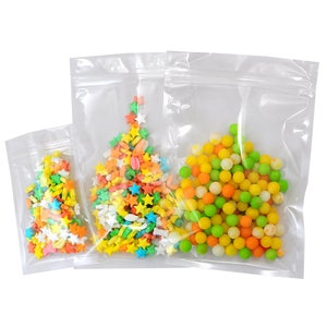 May include: Three clear plastic bags with zipper closures, containing colorful candy sprinkles and round candies. The bags are arranged in a triangular shape, with the largest bag at the top and the smallest bag at the bottom.