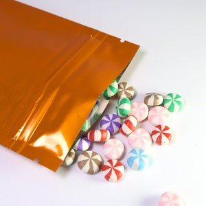 May include: A close-up of a bright orange metallic stand-up pouch with a zipper closure. The pouch is partially open, spilling out colorful round candies with a swirl design.