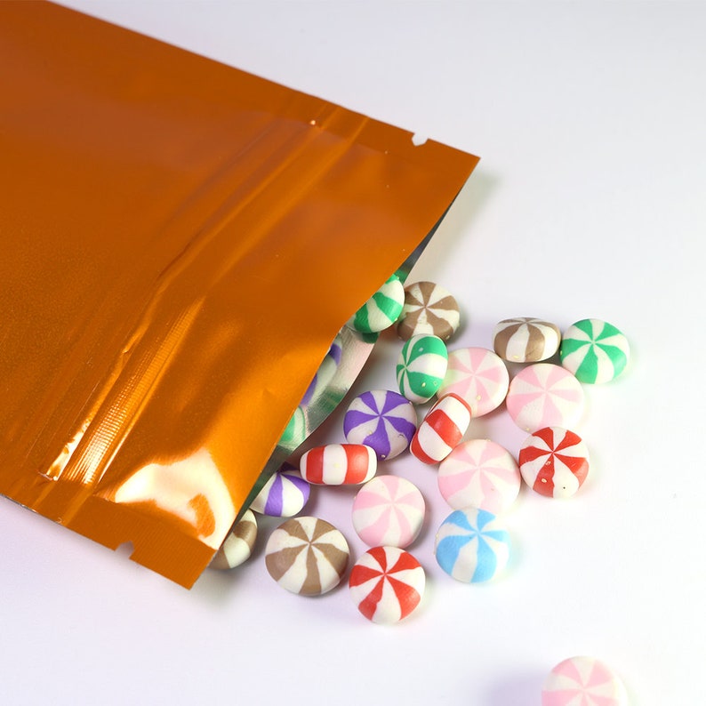 May include: A close-up of a bright orange metallic stand-up pouch with a zipper closure. The pouch is partially open, spilling out colorful round candies with a swirl design.
