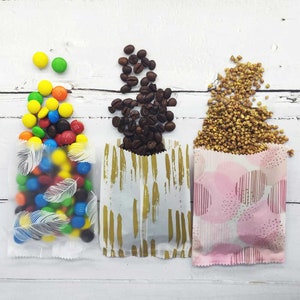May include: Three small paper bags with different patterns. The bag on the left is clear with white feather designs and filled with colorful M&Ms. The middle bag has a white background with gold brushstrokes and is filled with coffee beans. The bag on the right has a pink background with white polka dots and is filled with buckwheat groats.