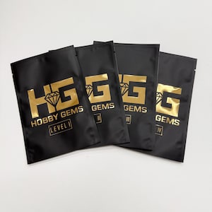 May include: Four black pouches with gold lettering and logos. The pouches are labelled "HOBBY GEMS" and "LEVEL I", "GEMS II", "GEMS III", and "GEMS IV". The pouches are sealed and appear to contain small items.