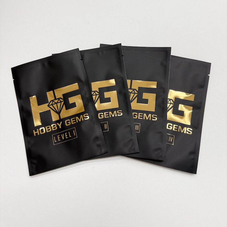 May include: Four black pouches with gold lettering and logos. The pouches are labelled "HOBBY GEMS" and "LEVEL I", "GEMS II", "GEMS III", and "GEMS IV". The pouches are sealed and appear to contain small items.