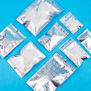 May include: An assortment of clear, silver, and blue iridescent zipper bags arranged on a bright blue background. The bags have a swirl pattern and a small hole at the top. The bags are empty and ready to be filled.