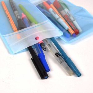 Op de afbeelding: Een doorzichtige plastic zak met een ritssluiting en een gat aan de bovenkant, gevuld met verschillende pennen en stiften, waaronder zwart, blauw, rood, groen en oranje. De zak ligt op een wit oppervlak.