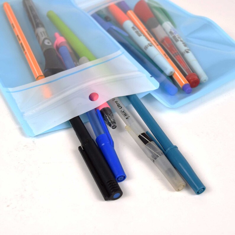 Op de afbeelding: Een doorzichtige plastic zak met een ritssluiting en een gat aan de bovenkant, gevuld met verschillende pennen en stiften, waaronder zwart, blauw, rood, groen en oranje. De zak ligt op een wit oppervlak.