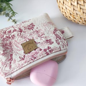 Könnte beinhalten: Kleine Reißverschlusstasche mit floralem Muster in Rosa und Weiß. Die Tasche hat ein Lederetikett mit einem Herzsymbol und ein rosa Etui ist teilweise sichtbar. Der Reißverschluss ist roségoldfarben.