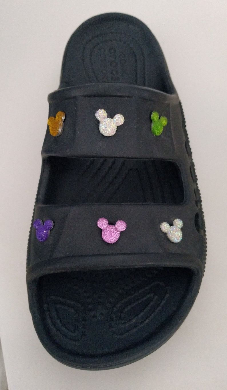May include: Black Crocs sandals with a double strap design. The sandals have five Mickey Mouse charms attached to them, each with a different color of glitter.