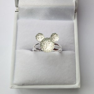 K&ouml;nnte beinhalten: Ein silberner Ring mit einem Mickey-Maus-Kopf-Design. Der Kopf besteht aus kleinen, funkelnden Steinen.