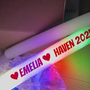 May include: A white LED foam glow stick with the text "EMELIA" and "HAVEN 2023" in red, along with red heart symbols. The stick is illuminated with a rainbow of colors.