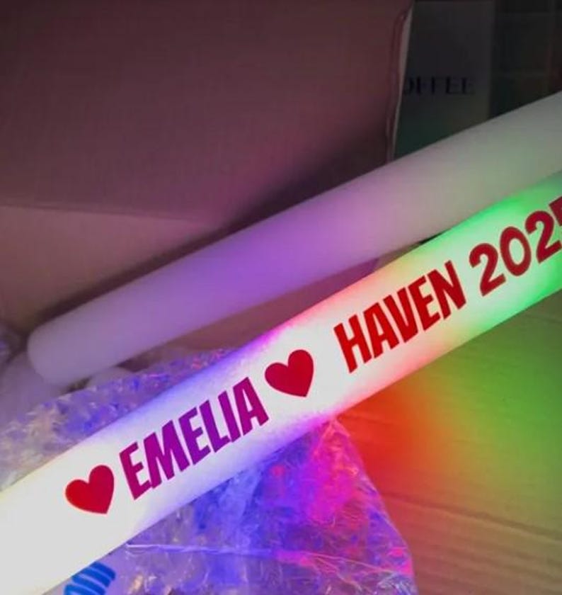 May include: A white LED foam glow stick with the text "EMELIA" and "HAVEN 2023" in red, along with red heart symbols. The stick is illuminated with a rainbow of colors.
