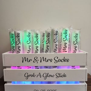 May include: A white wooden crate with the text "Mr & Mrs Sacks" and "Grab A Glow Stick". The crate holds several glowing tubes labeled "Mrs Sacks". The date "04-08-2025" is printed on the front of the crate.