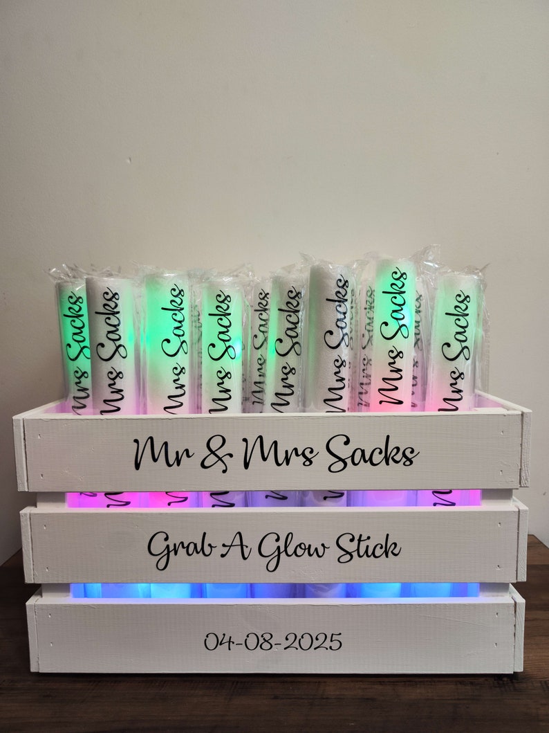 May include: A white wooden crate with the text "Mr & Mrs Sacks" and "Grab A Glow Stick". The crate holds several glowing tubes labeled "Mrs Sacks". The date "04-08-2025" is printed on the front of the crate.