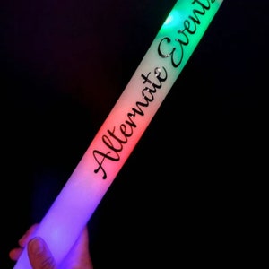 May include: A glowing foam stick with the text "Alternate Events" in black script. The stick transitions from purple to red to green, with the text running along the length of the stick. The background is black.