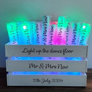 May include: A white wooden crate with multi-colored LED lights, holding glowing light sticks. The crate has the text "Light up the dance floor", "Mr & Mrs New", and "12th July 2024". The light sticks are labeled "Mrs New".